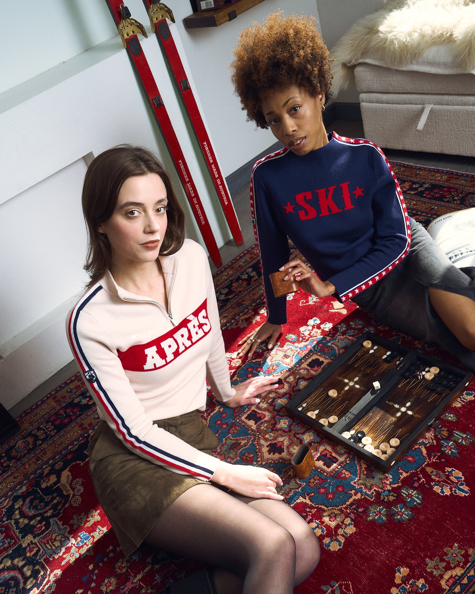 Two women playing backgammon on a persian rug with 'APRES' and 'Ski' text on their sweaters.